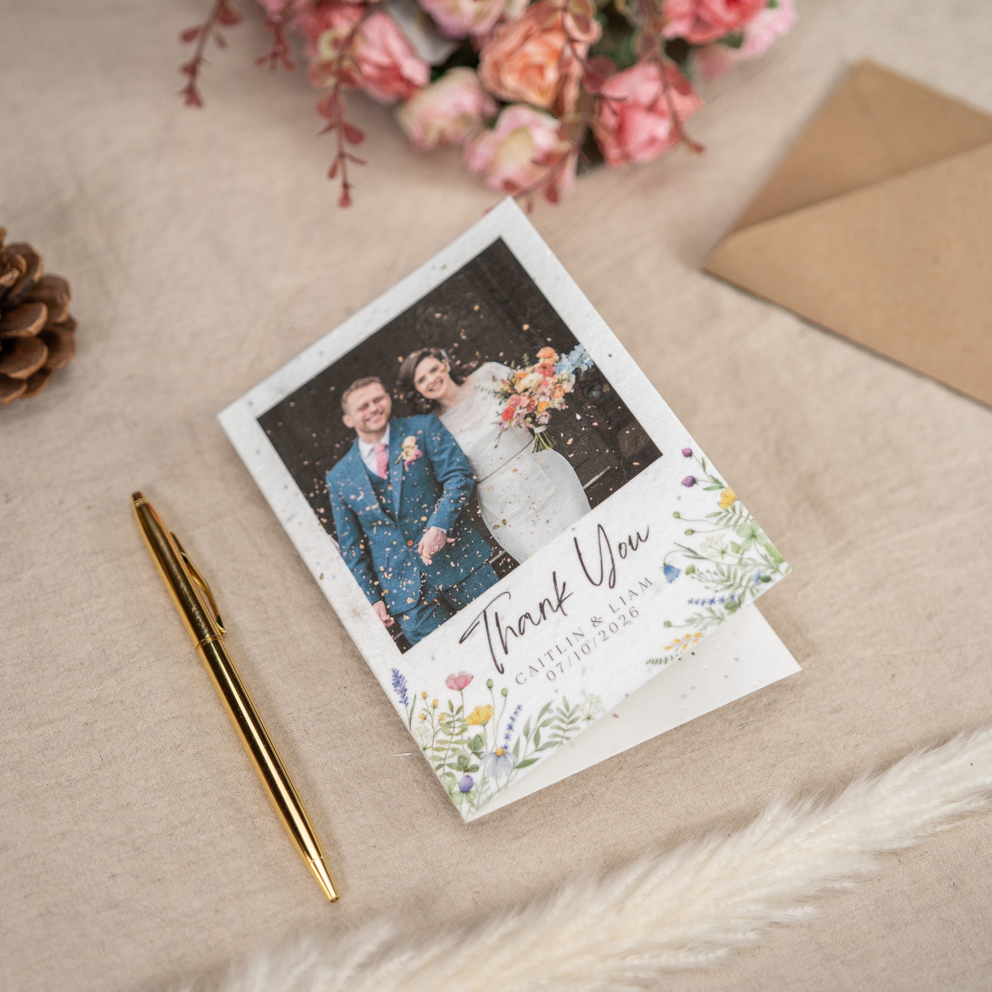 On a linen lined table sits a Wedding Thank You Card. On the cover of the thank you card is a picture of a newly married couple walking through confetti, underneath the photograph is customisable text - in this instance being the names of the couple and the wedding date.