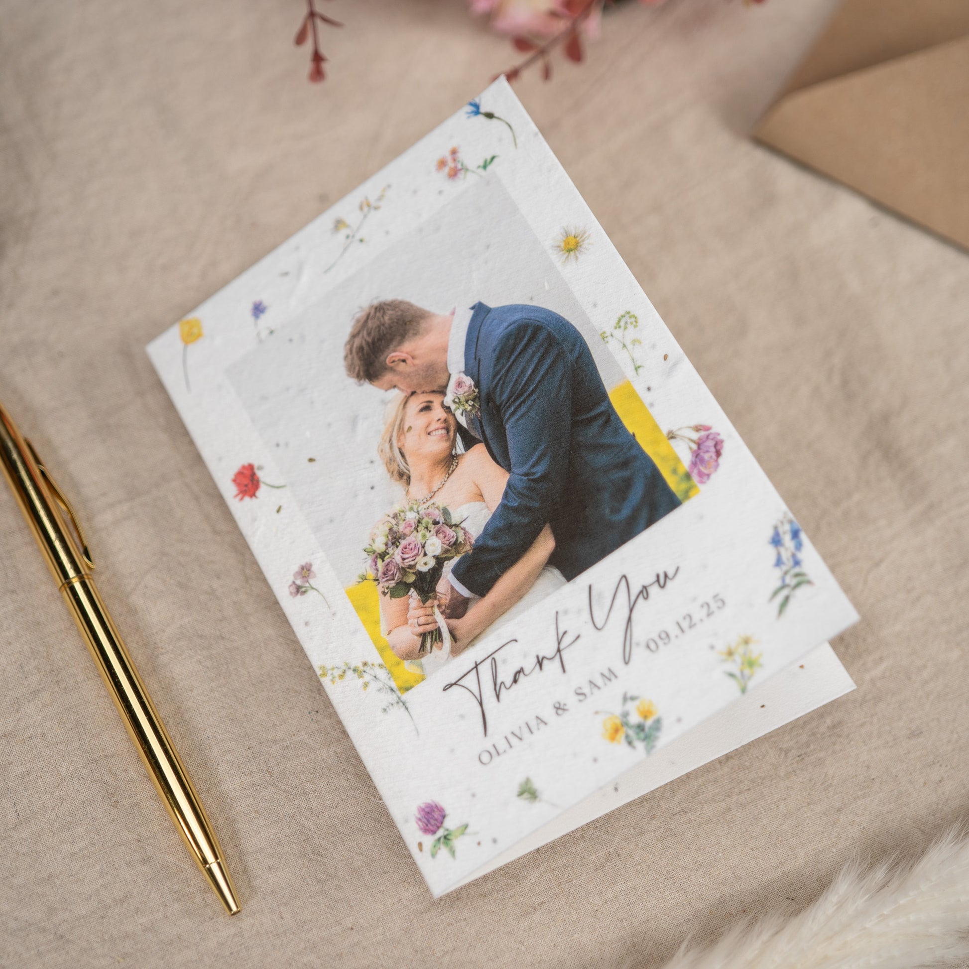 On a linen lined table sits a Wedding Thank You Card. On the cover of the thank you card is a picture of a newly married couple walking through confetti, underneath the photograph is customisable text - in this instance being the names of the couple and the wedding date.
