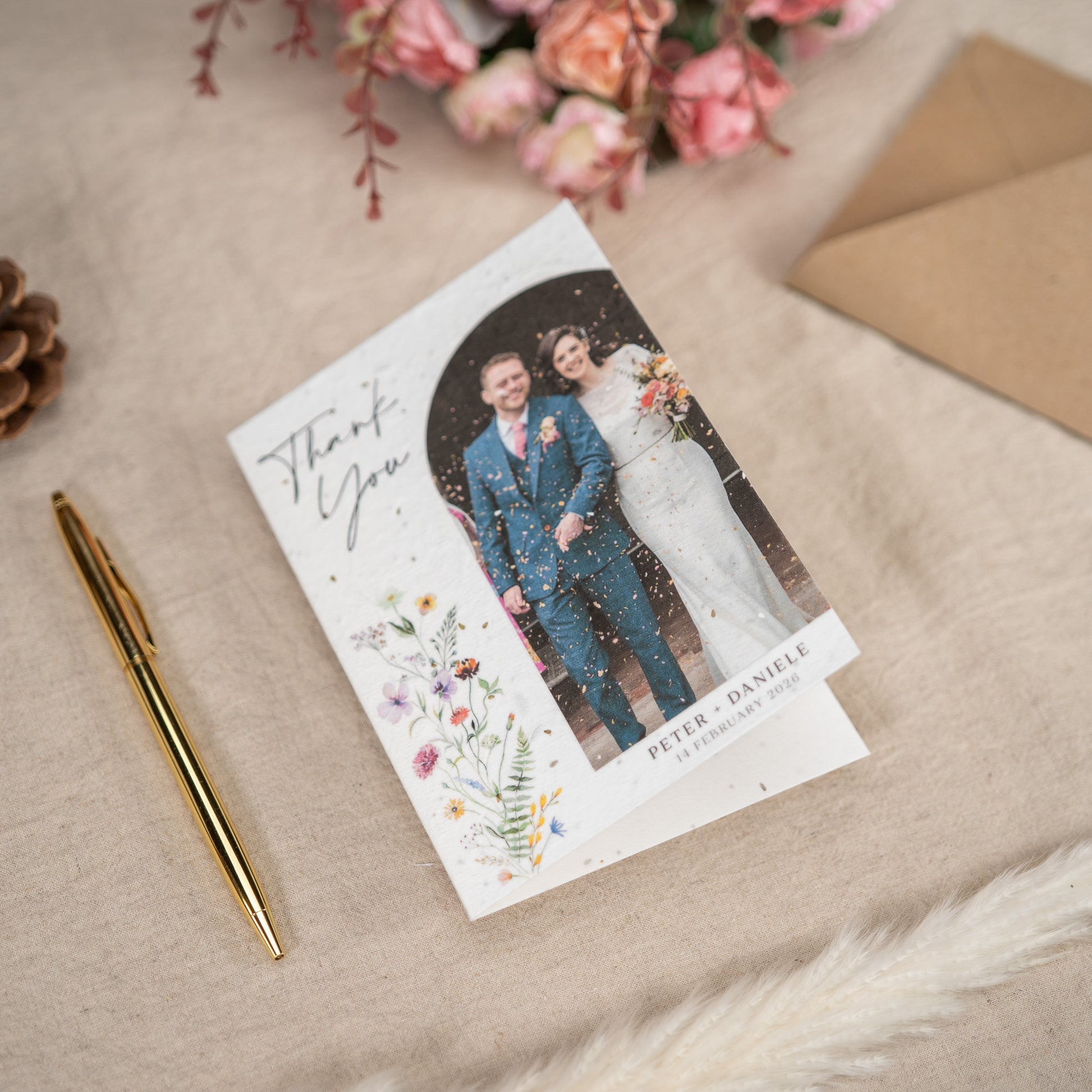 Wedding thank you card with a photo of a couple, surrounded by flowers and an envelope on a textured surface.