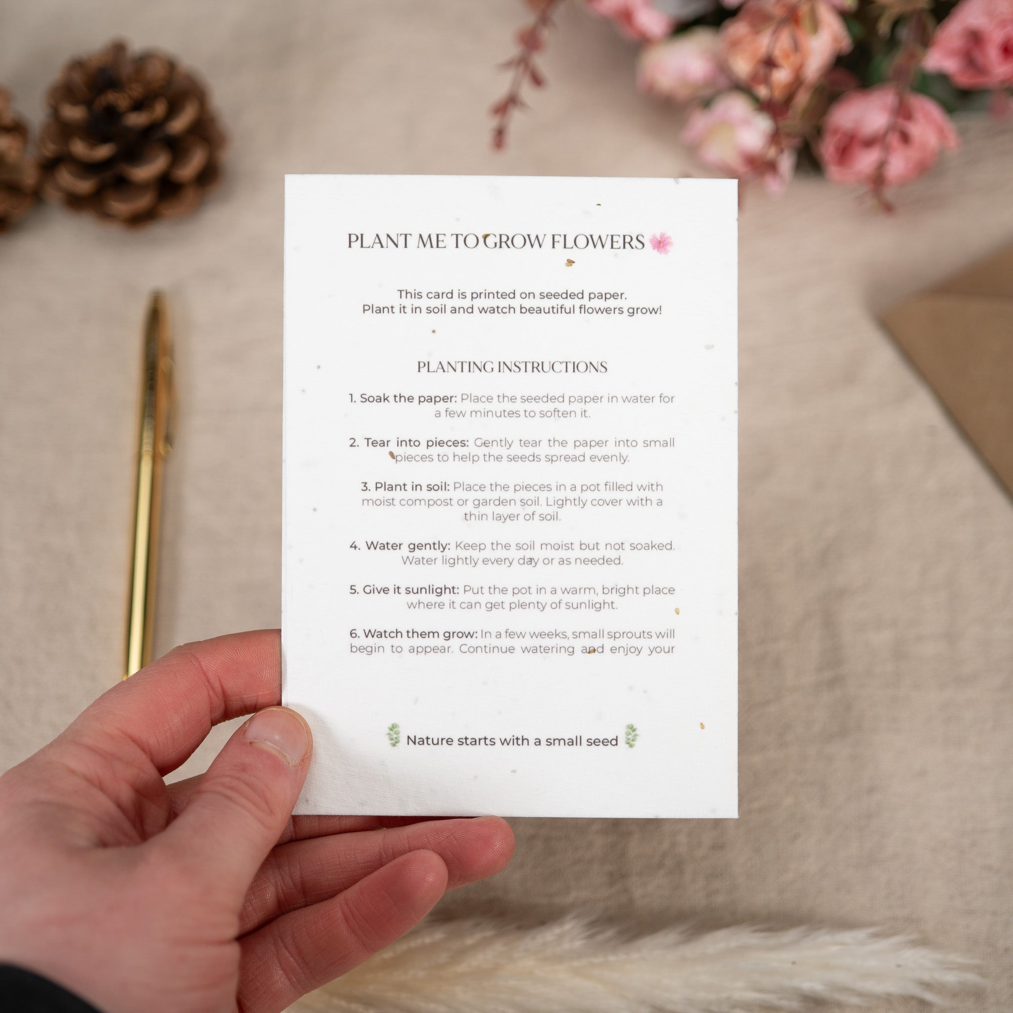 Hand holding a white card with planting instructions for seeded paper, surrounded by decorative items on a beige surface.