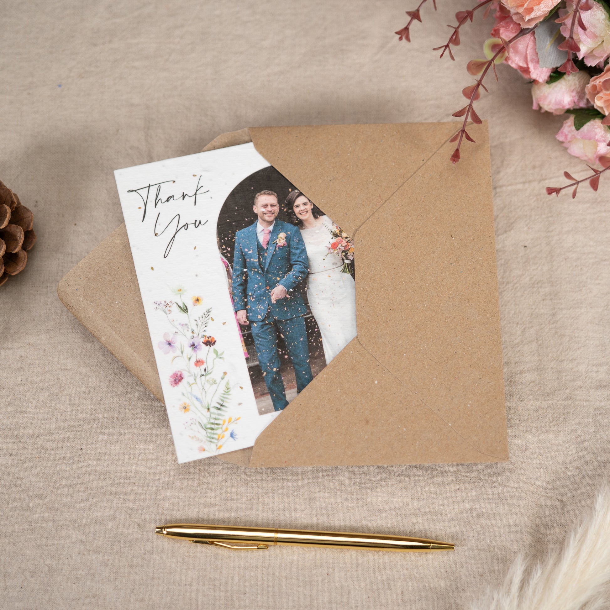 On a linen lined table sits a Wedding Thank You Card sticking out of a kraft brown envelope. On the cover of the thank you card is a picture of a newly married couple walking through confetti
