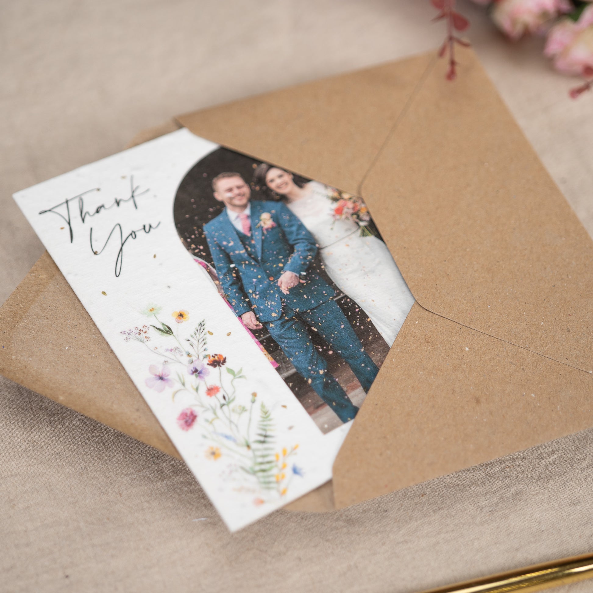 On a linen lined table sits a Wedding Thank You Card. On the cover of the thank you card is a picture of a newly married couple walking through confetti