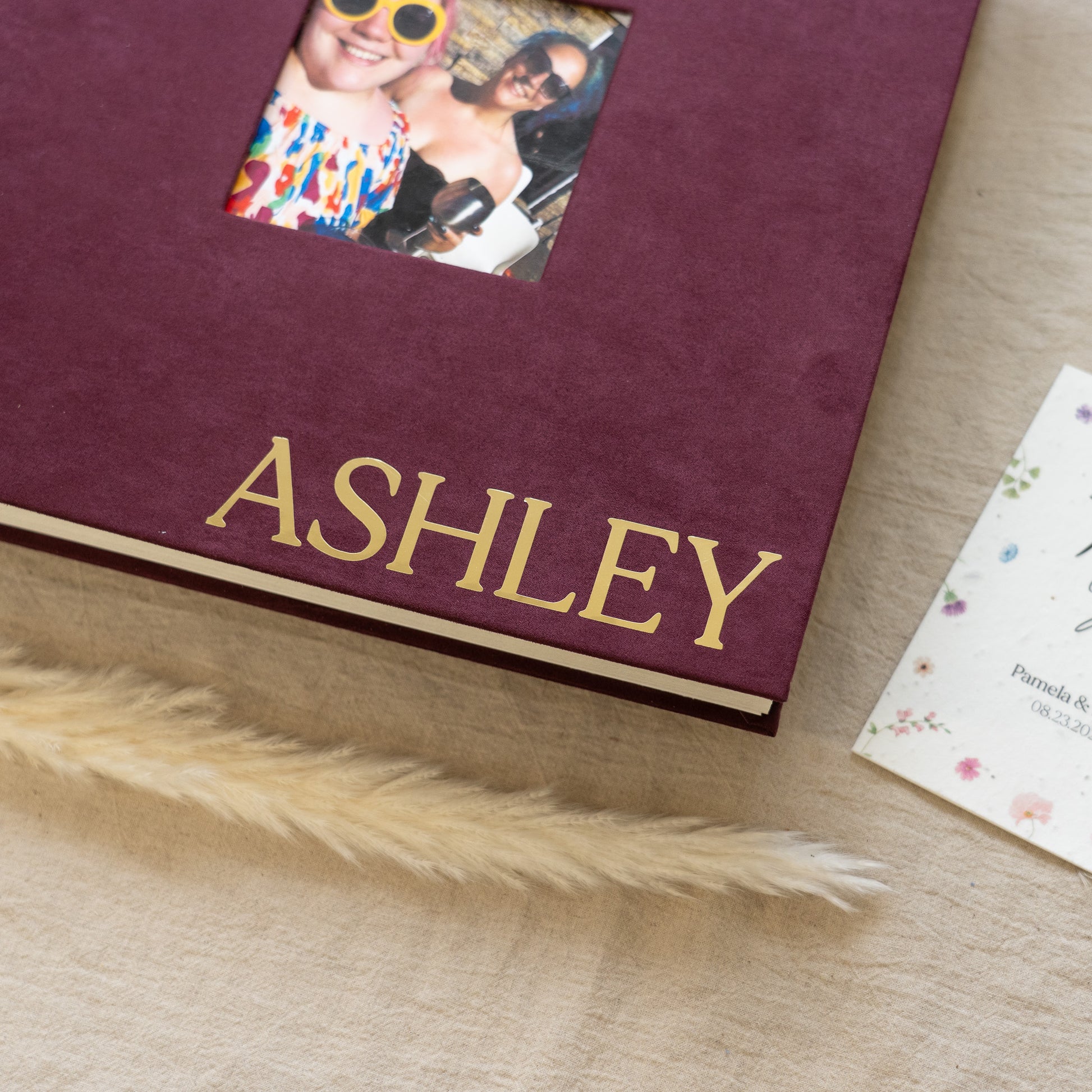 Maroon photo album with 'ASHLEY' on it, featuring a small window with photos on a wooden surface.