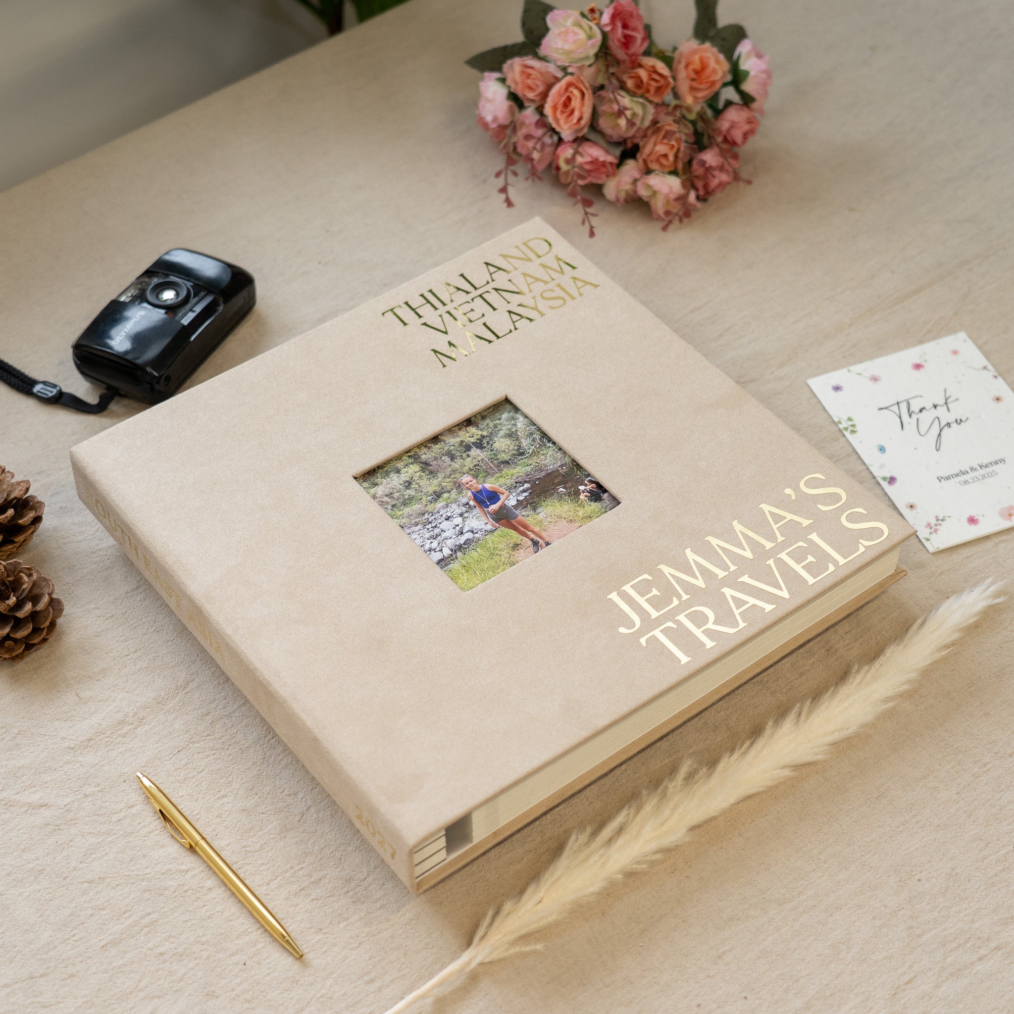 Travel photo album with 'Jemma's Travels' on a table with a camera and flowers.