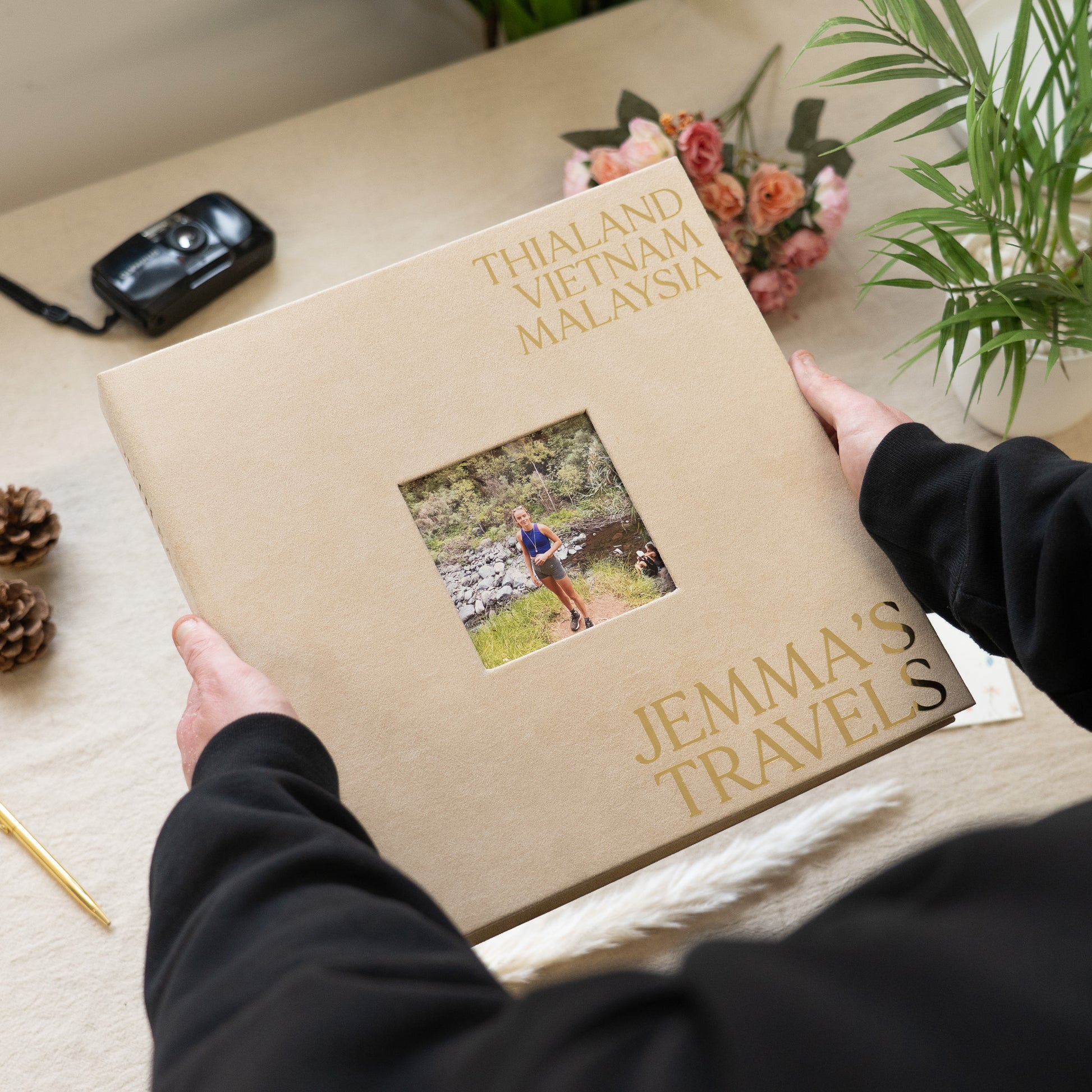 Album titled 'Jemma's Travels' with a photo of a person holding a camera, placed on a surface with a camera and flowers in the background.