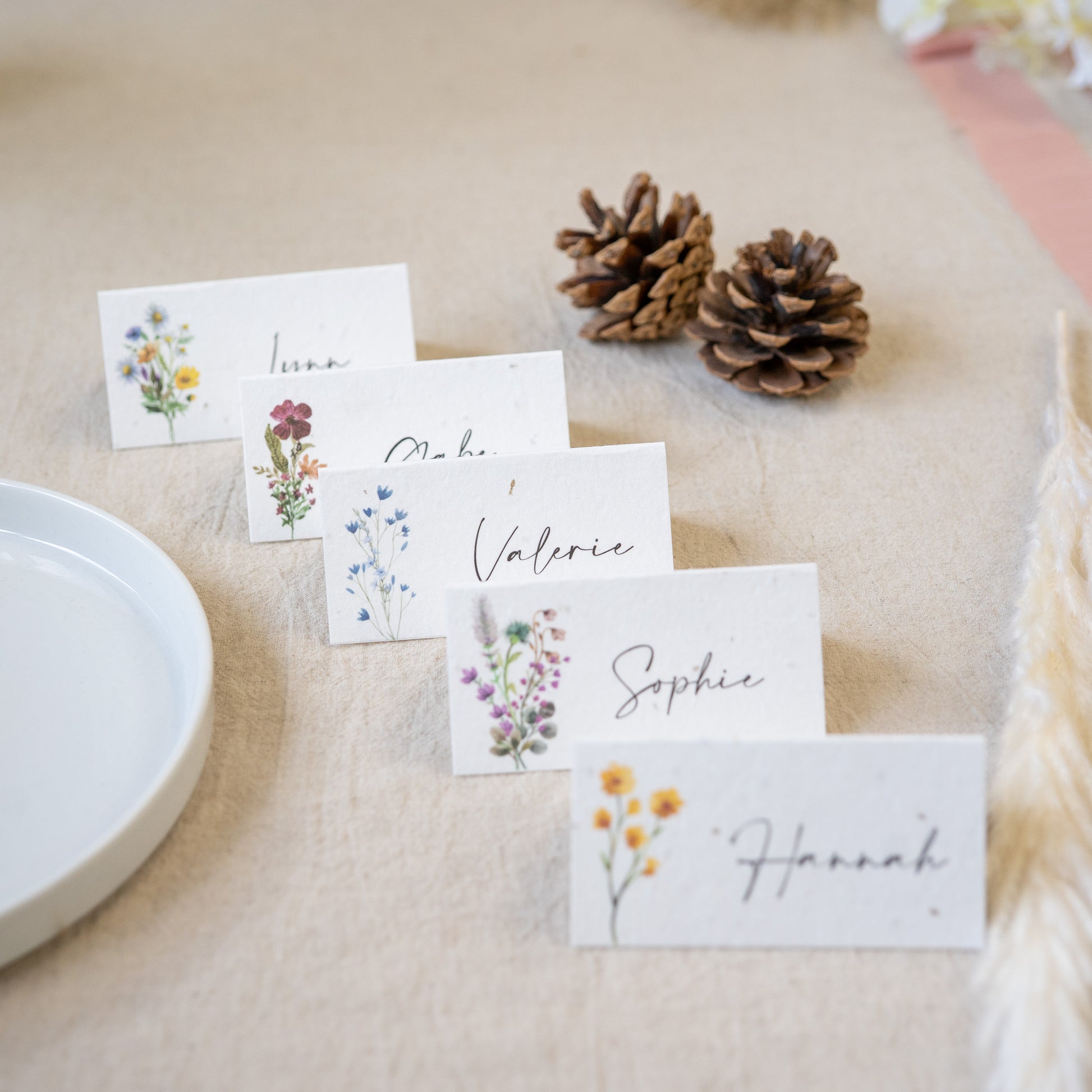 Place cards with floral designs and names on a table with pine cones.
