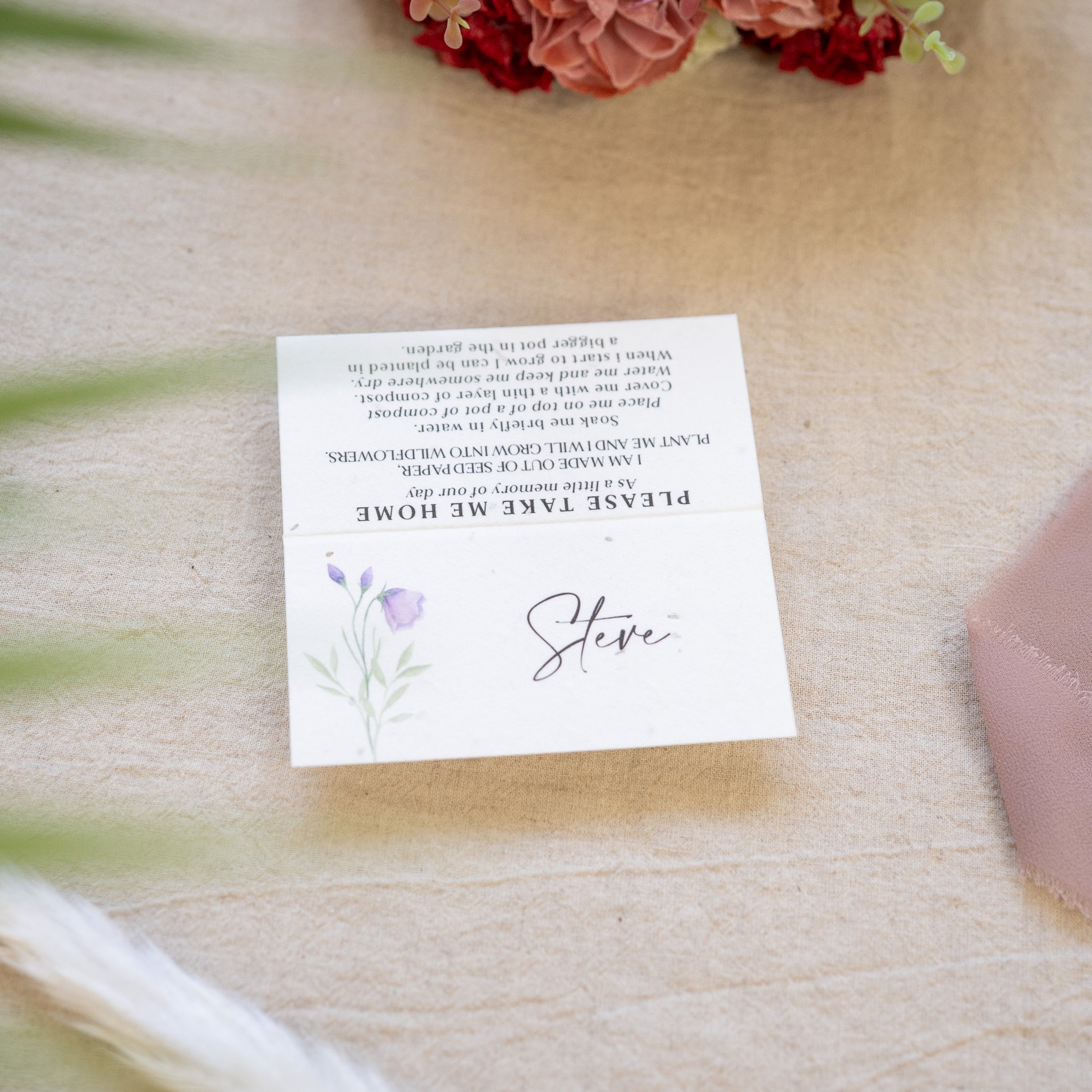 White card with text and floral design on a wooden surface with flowers.