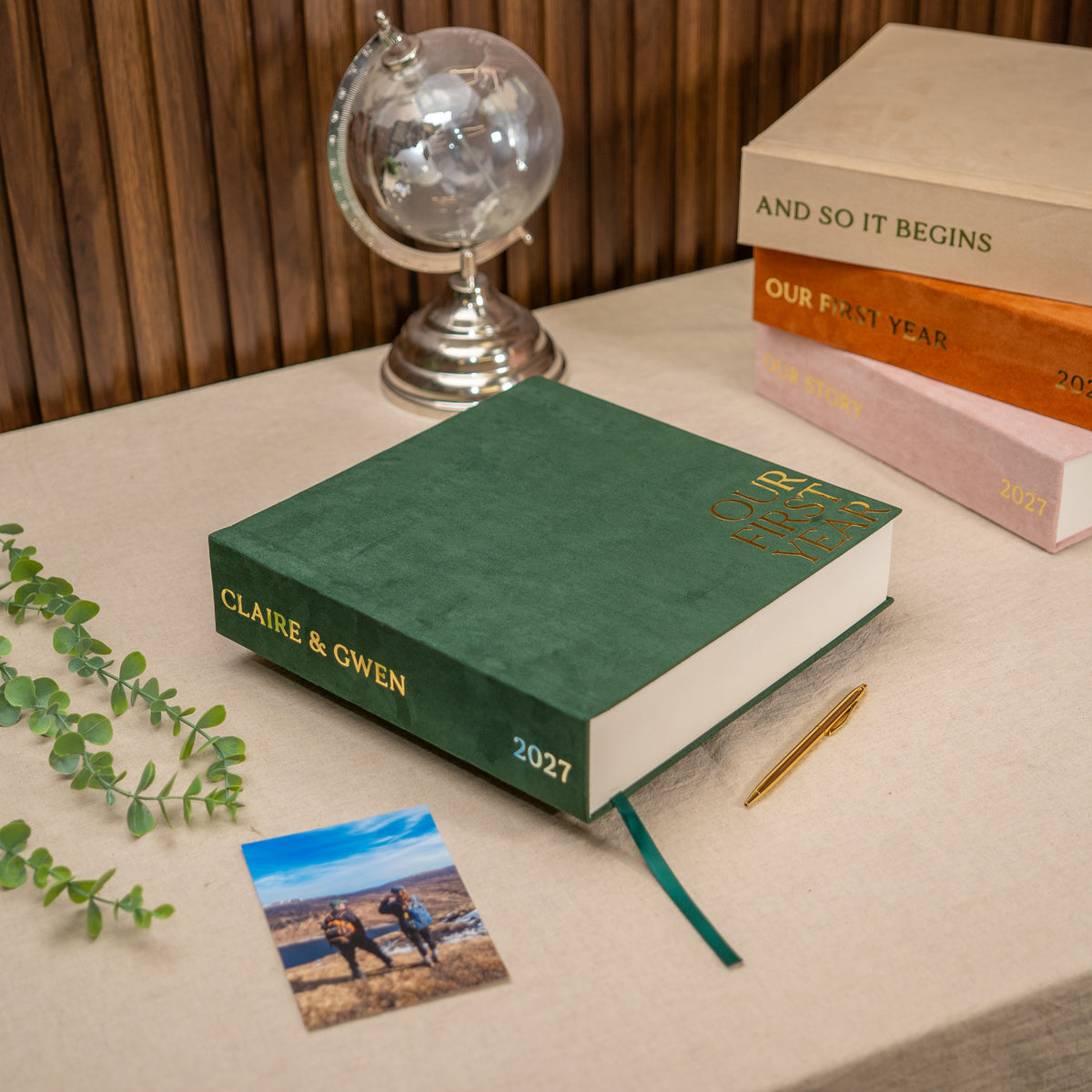 A green hardcover photo book titled 'Our Love Story 2027' lies on a beige tabletop, styled with a small photo, gold pen, books, and a globe against a wood-paneled background.