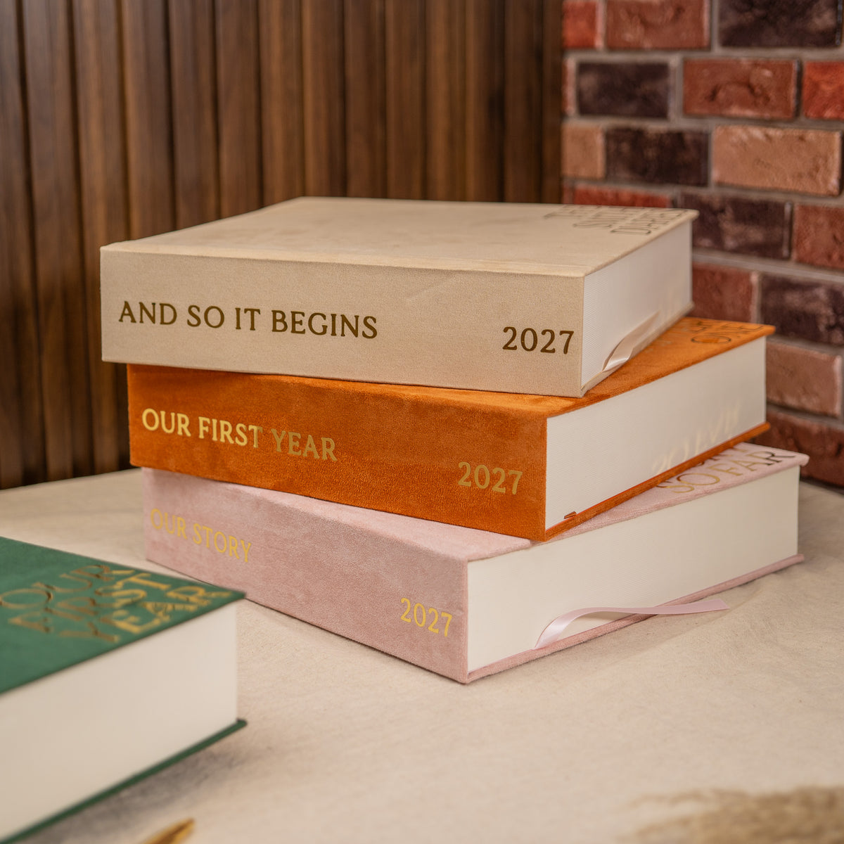 A stack of four hardcover books with gold foil titles including 'AND SO IT BEGINS', 'OUR FIRST YEAR', and '2027' on a tabletop, with a brick wall and wood paneling in the background.