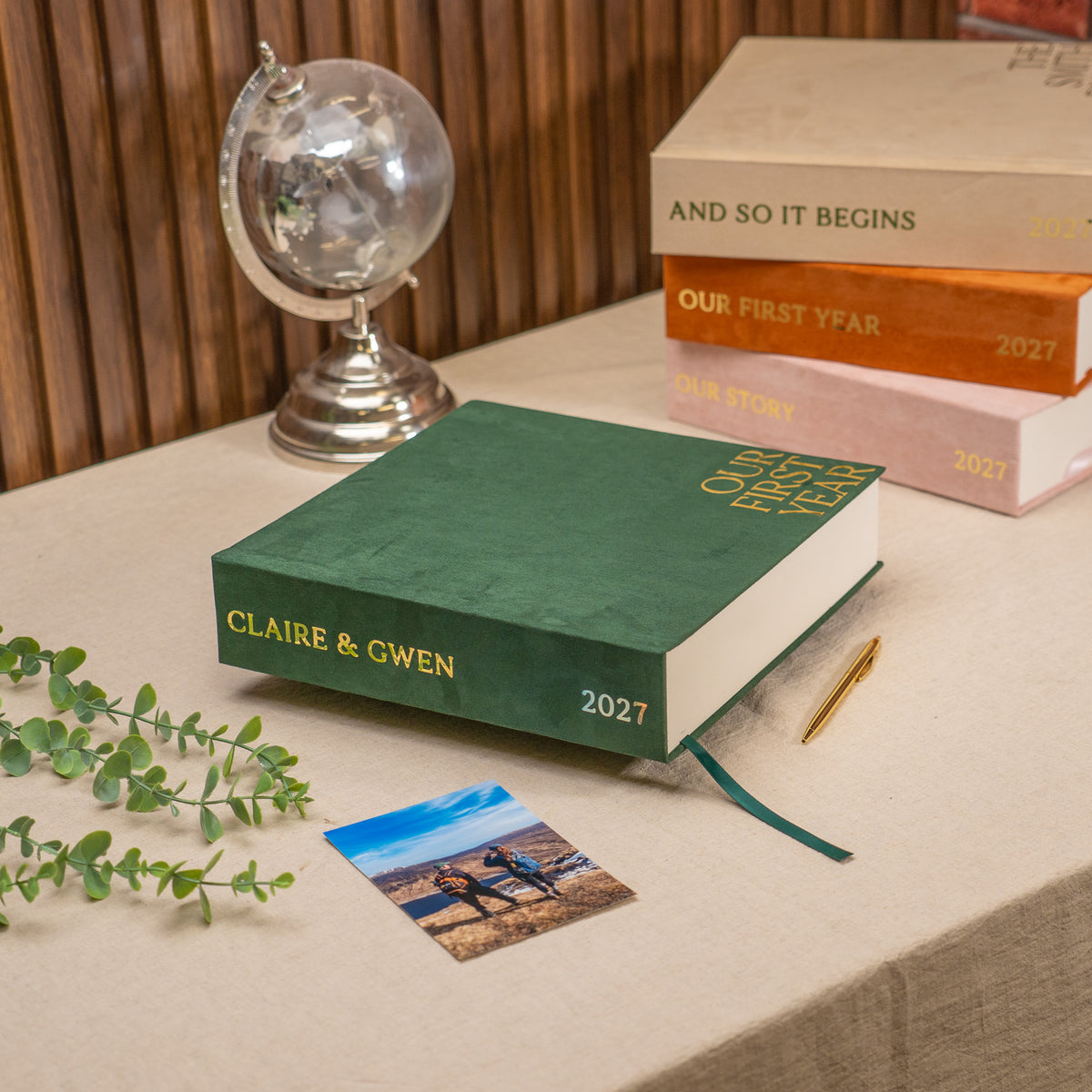 A personalized hardcover wedding calendar or photo book for 'Claire & Gwen 2027' in deep green with gold lettering, styled on a beige tablecloth with a small globe, books, a pen, and a photo print.