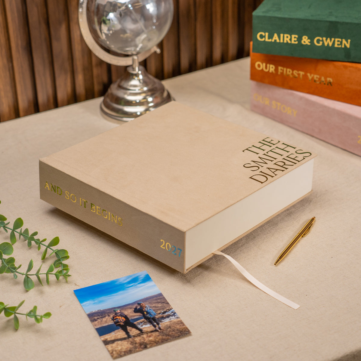 A beige hardcover journal titled 'THE SMITH DIARIES' sits on a tabletop with a white ribbon bookmark extending from the page. The scene includes a small printed photo card, a gold pen, greenery, a silver globe, and stacked books in the background.