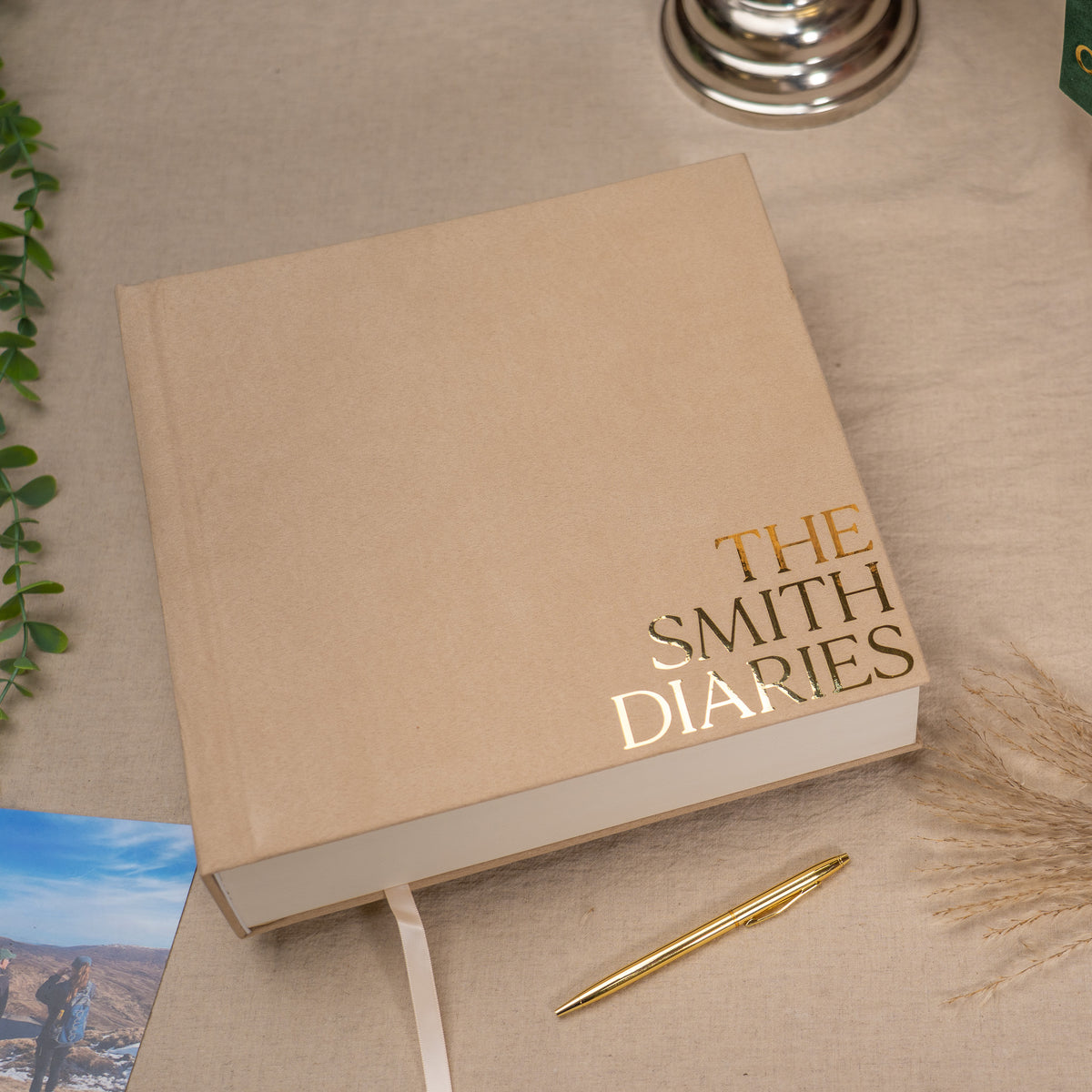 A beige hardcover journal titled 'THE SMITH DIARIES' in gold foil lettering, styled on a linen surface with a gold pen, dried grass, and a small photo card.
