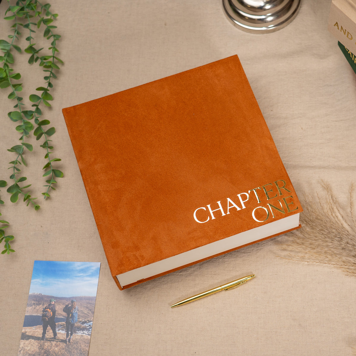 A custom hardcover book or photo album styled on a beige linen surface, featuring a textured orange cover with 'CHAPTER ONE' printed in gold foil. The book is surrounded by lifestyle props including a green eucalyptus sprig, a gold pen, a small printed photo, and dried grass.