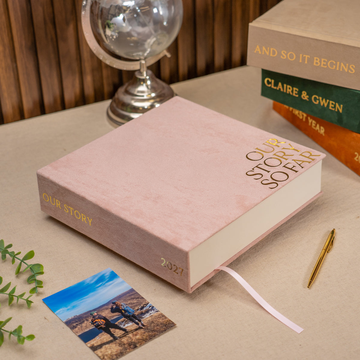 A pink hardcover book titled 'Our Story So Far 2027' styled on a beige tabletop with a ribbon bookmark, alongside a small photo card, gold pen, books, and a globe in the background.
