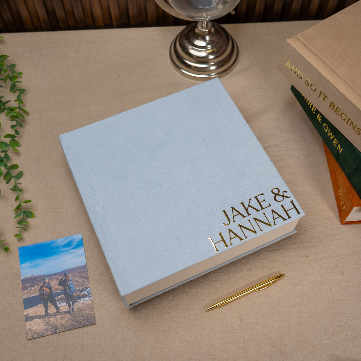 A personalized hardcover photo book with a light blue cover and gold foil text reading 'JAKE & HANNAH', styled on a beige fabric surface with a gold pen, a small photo print, and other decorative props.