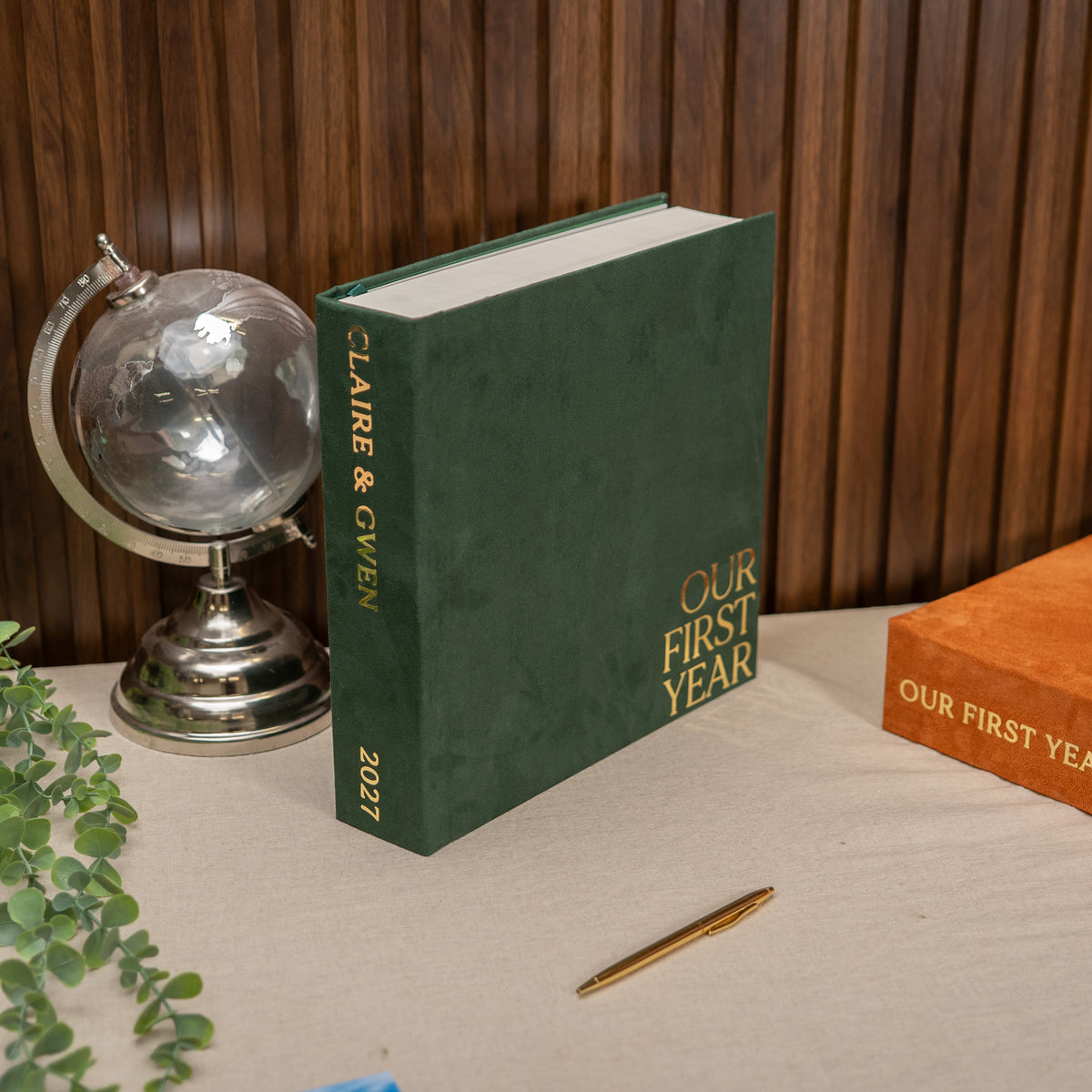 A hardcover book titled 'Our First Year 2027' by Claire & Gwen stands open on a tabletop, flanked by a silver globe and a gold pen, with another matching book partially visible on the right against a wood-paneled background.