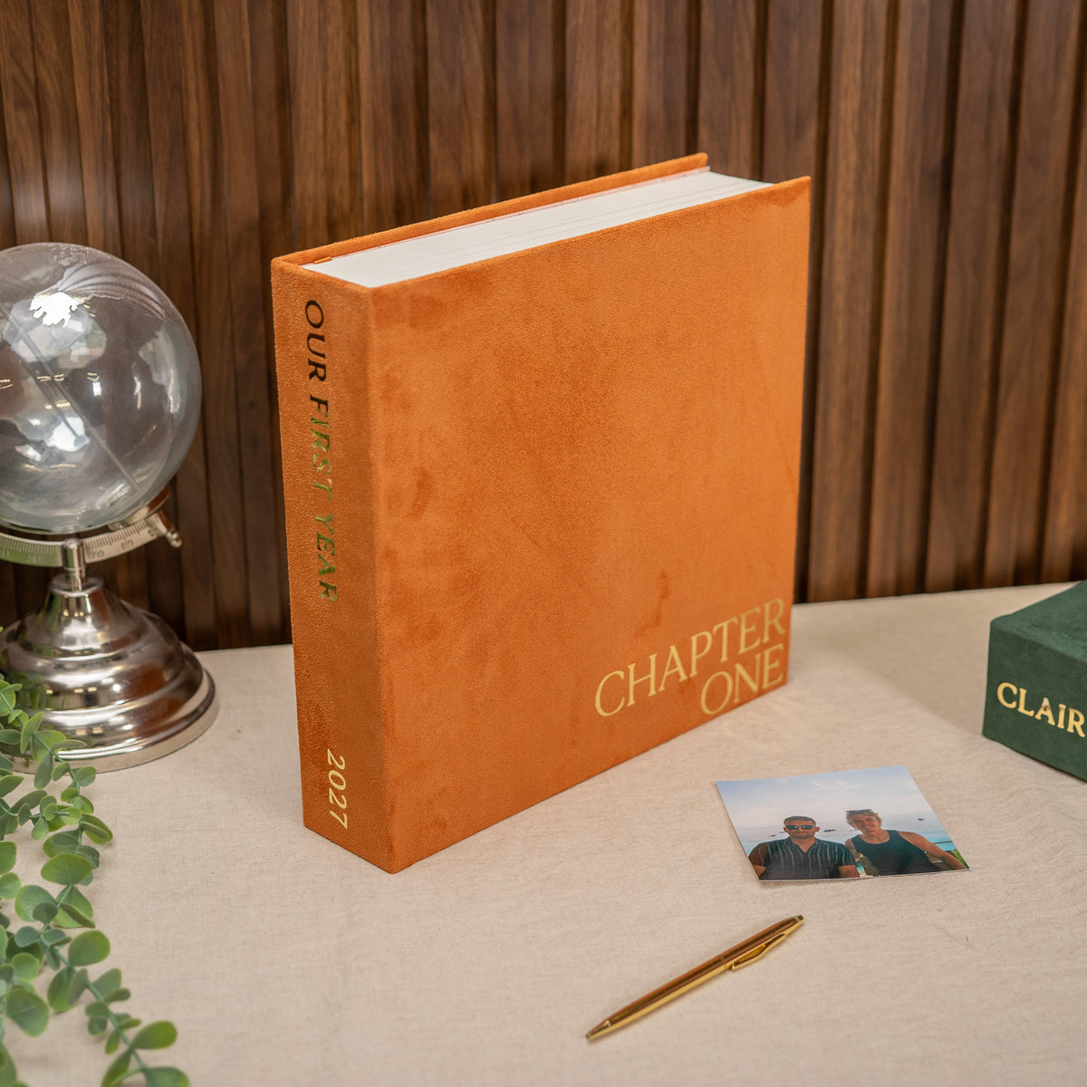 An orange hardcover book standing upright on a beige tabletop, with the title 'CHAPTER ONE' and '2027' printed on the spine and cover. A small photo card, gold pen, and green box are nearby, with a wood-paneled wall in the background.