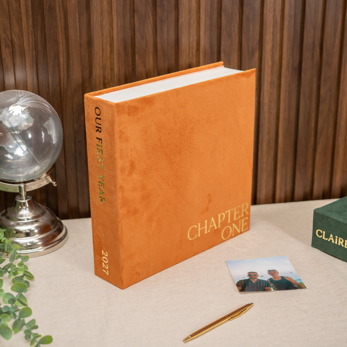 An orange hardcover book standing upright on a tabletop, with the spine reading 'OUR FIRST 30 YEARS 2027' and the front cover reading 'CHAPTER ONE.' The scene includes a wood-paneled wall background, a silver globe, greenery, a pen, a small photo, and part of another green box.