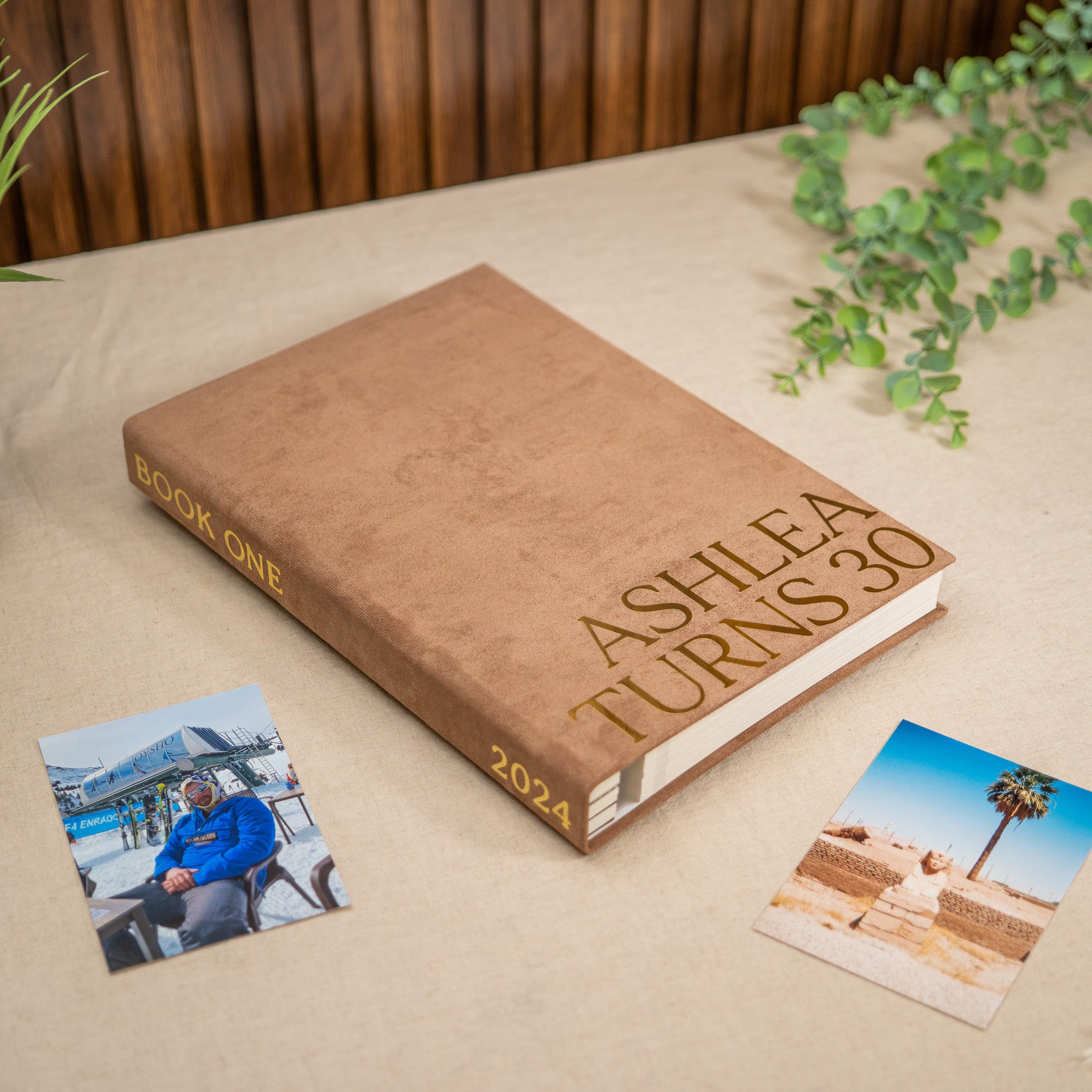 A brown hardcover photo book titled 'ASHLEA TURNS 30' with 'BOOK ONE' and '2024' on the spine, styled on a beige fabric surface with two printed photos and green trailing plants.
