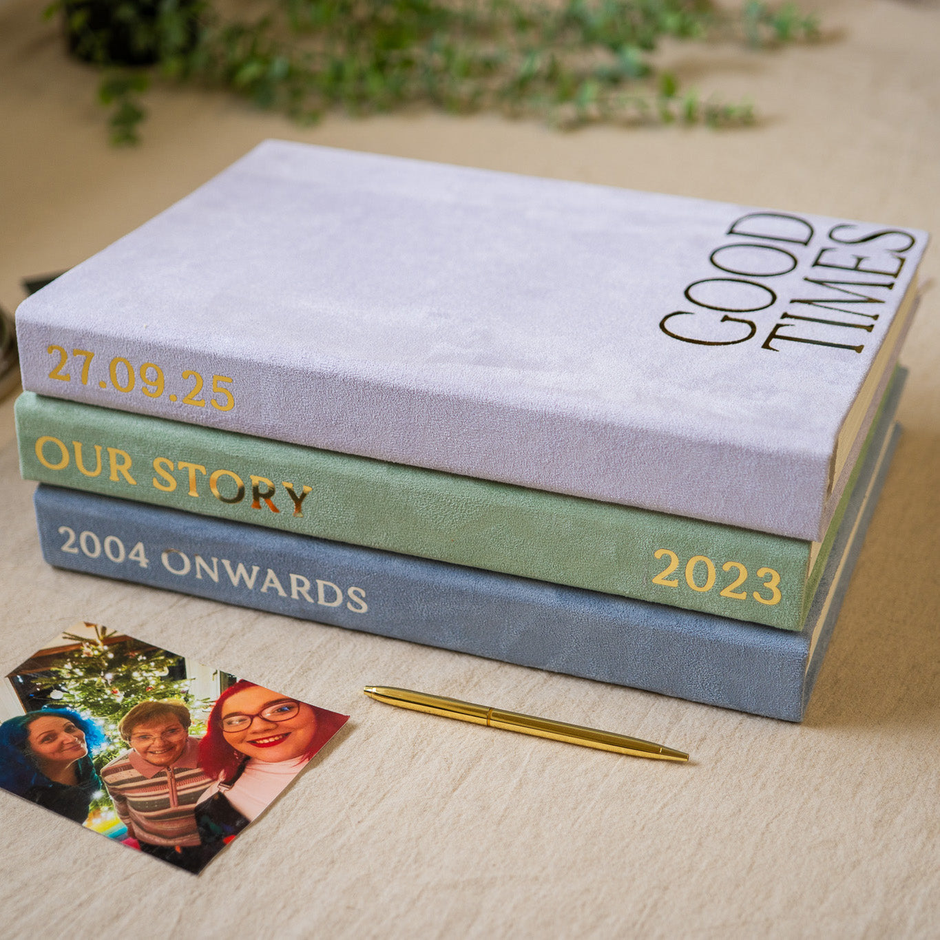 Three personalized hardcover books stacked on a light wood surface, with small printed photos and a gold pen nearby. The spines read 'GOOD TIMES', 'OUR STORY', and '2004 ONWARDS' in gold lettering.