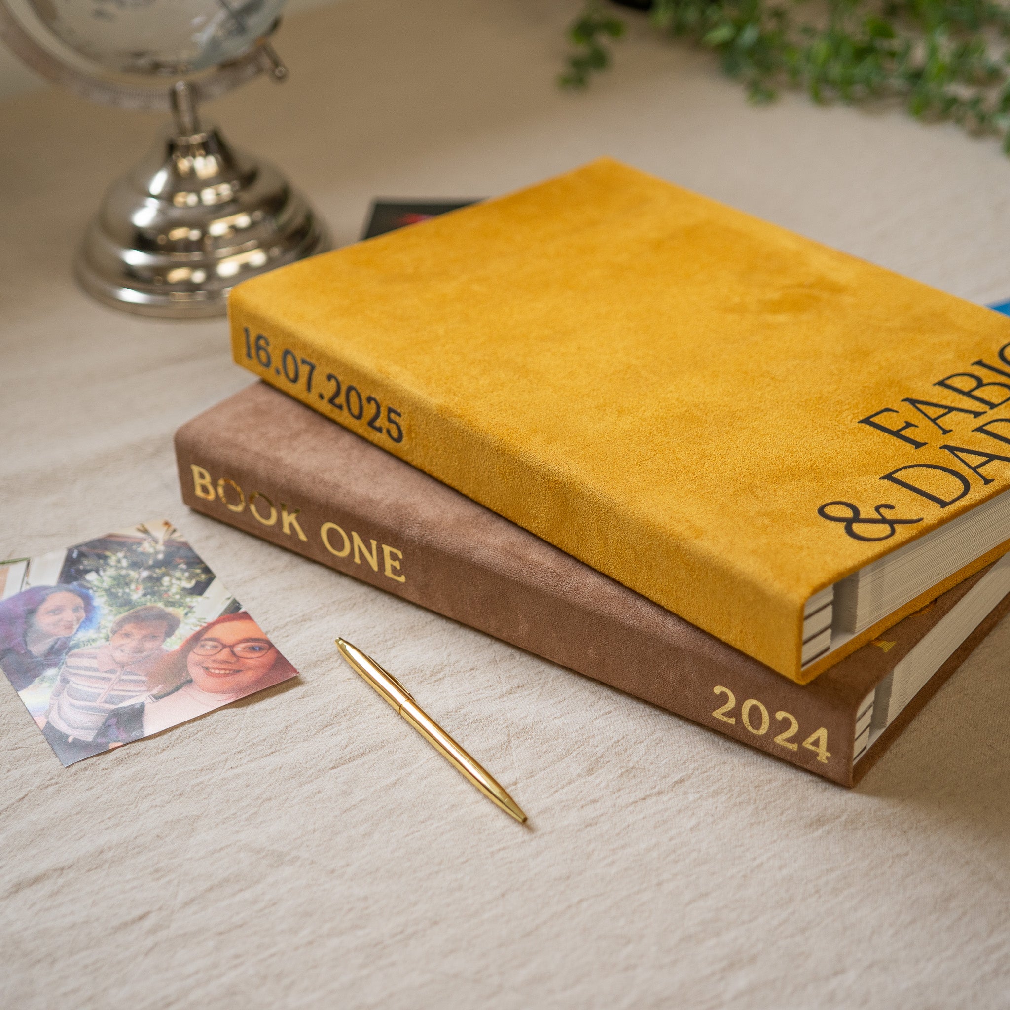 A personalized hardcover book with a mustard yellow velvet cover and gold foil lettering, stacked on top of a brown velvet book with gold foil lettering. A gold pen and small printed photos rest on a light beige fabric surface.