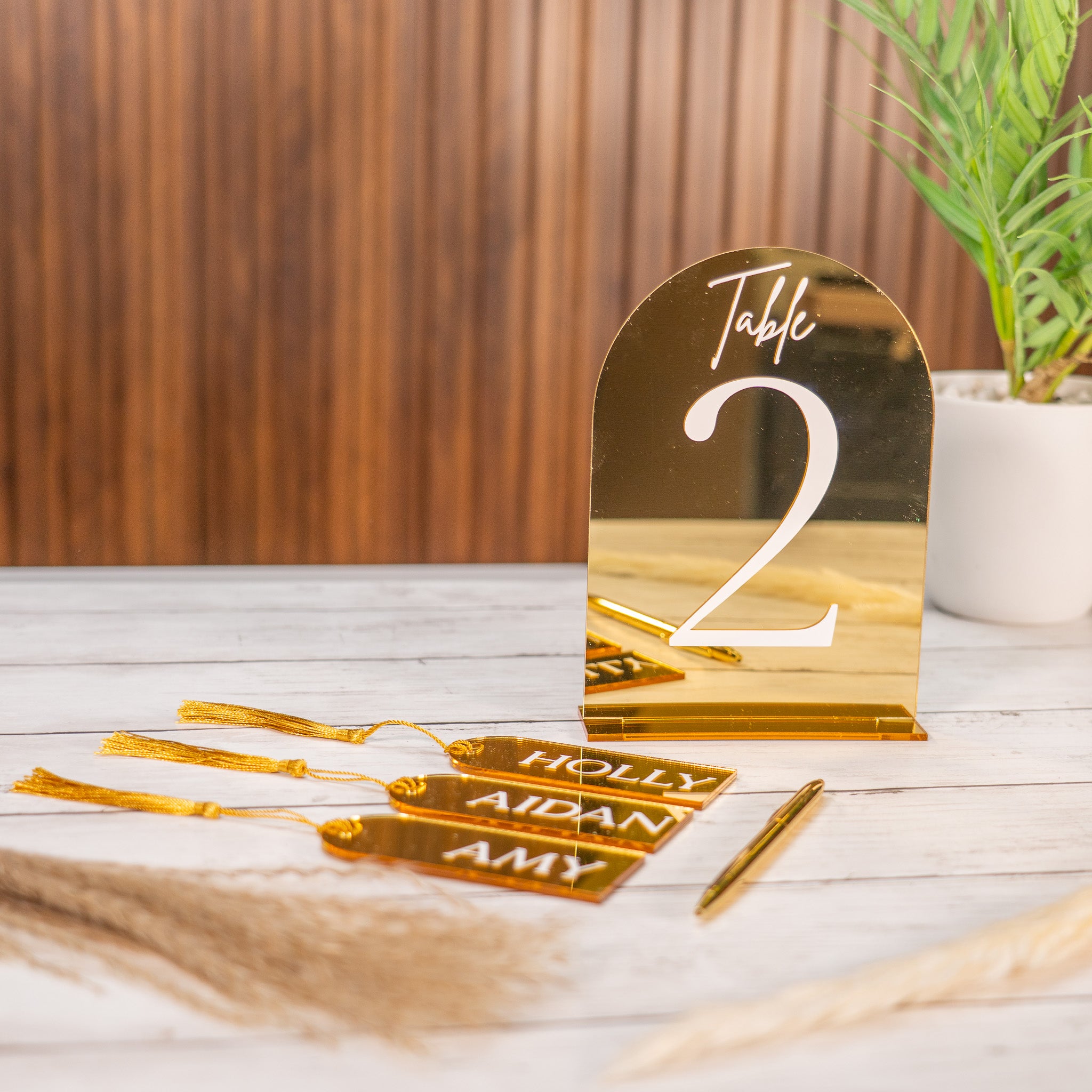 Gold table number and name tags on a wooden surface with a plant in the background