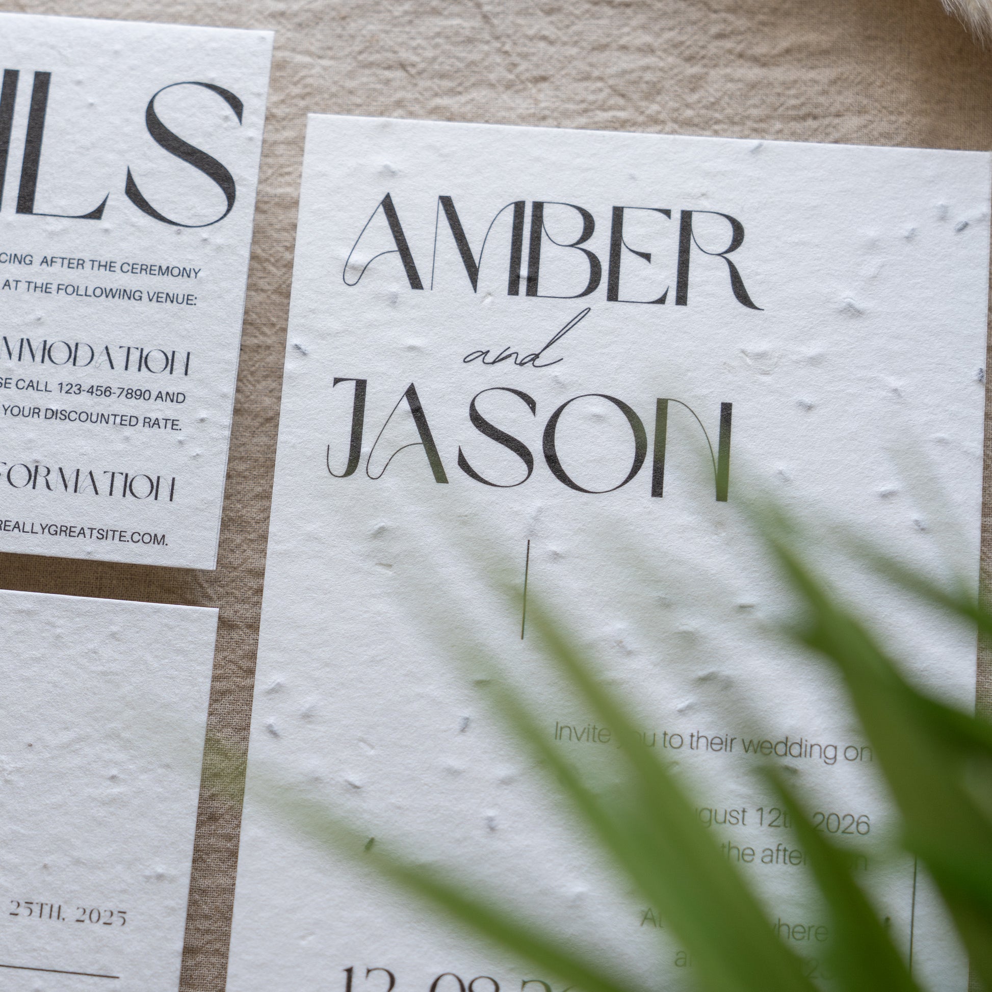 Wedding invitation card with 'Amber and Jason' on a textured surface with green leaves.