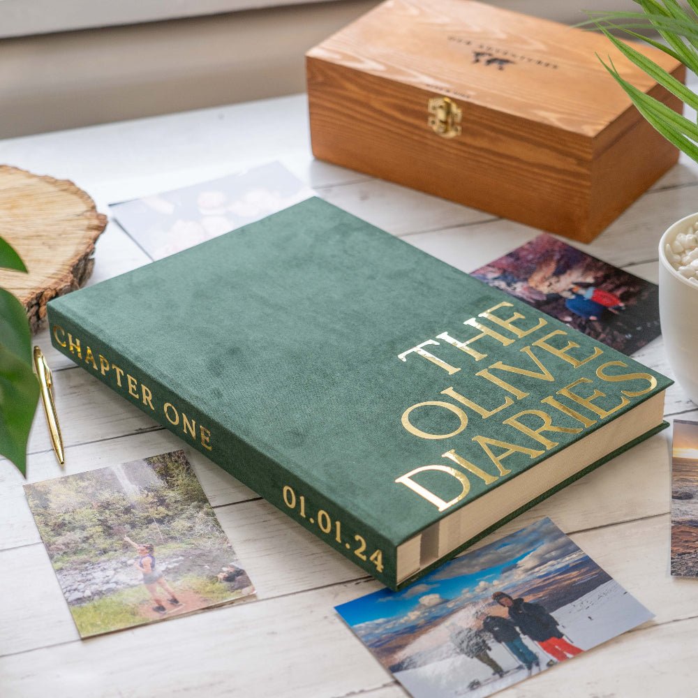 Green book titled 'The Olive Diaries' with gold text on a wooden surface with photos and a wooden box.