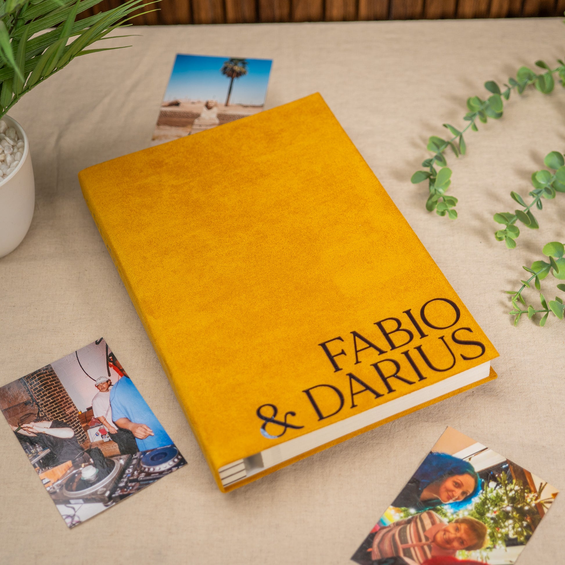 A hardcover book with a textured mustard yellow cover and the names 'FABIO & DARIUS' printed in black serif font on the front, styled on a beige fabric surface with scattered printed photos and green plant sprigs.