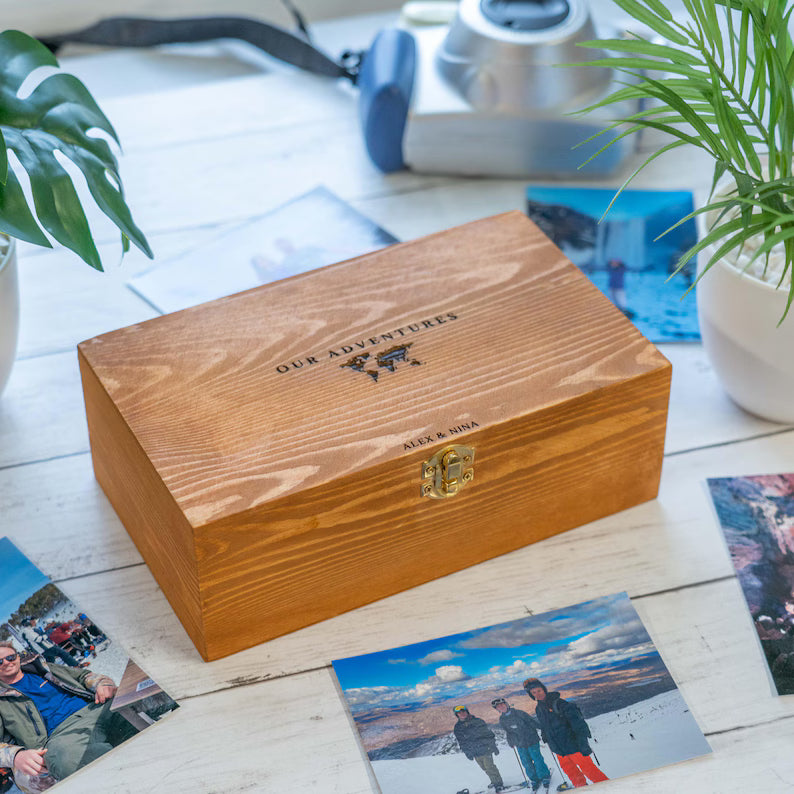 A rectangular wooden keepsake box with a hinged lid, engraved with 'OUR ADVENTURES' and a mountain graphic, with 'ALEX & MIA' below it. The box is styled on a white wooden table surrounded by printed photos, a potted plant, and a camera.