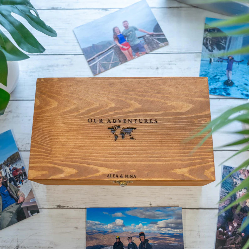 A rectangular wooden keepsake box with engraved text reading 'OUR ADVENTURES' and 'ALEX & NINA' sits on a white wood plank surface surrounded by scattered travel photos and green plant leaves.
