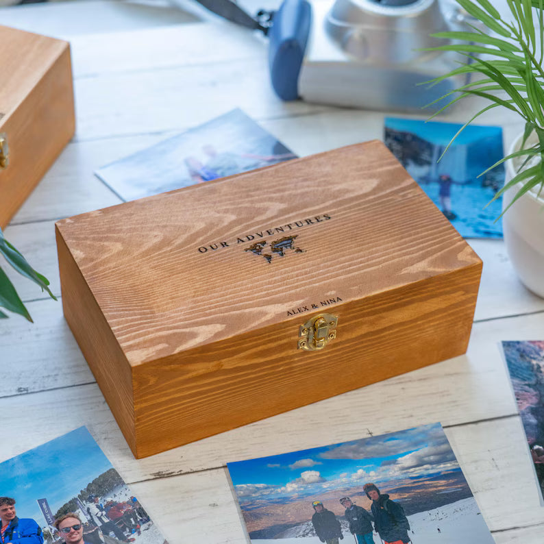 A personalized wooden photo box with a hinged lid engraved with 'OUR ADVENTURES' and 'MEL & HINA', shown on a white wooden tabletop with printed photos scattered around it.