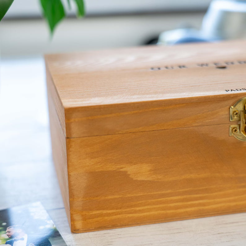 A close-up of a wooden keepsake box with a warm honey-brown finish, brass clasp, and partially visible embossed text on the lid, resting on a light surface.
