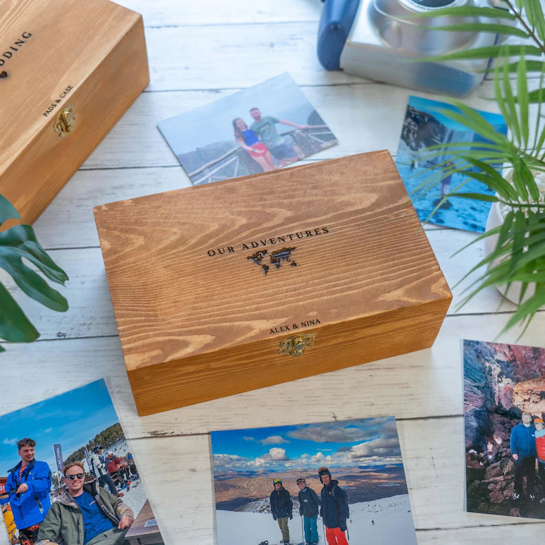 A wooden keepsake box with a custom engraved design reading 'OUR ADVENTURES' and 'ALEX & MIA' with a small mountain graphic, displayed on a white wooden surface surrounded by travel photos and green plants.