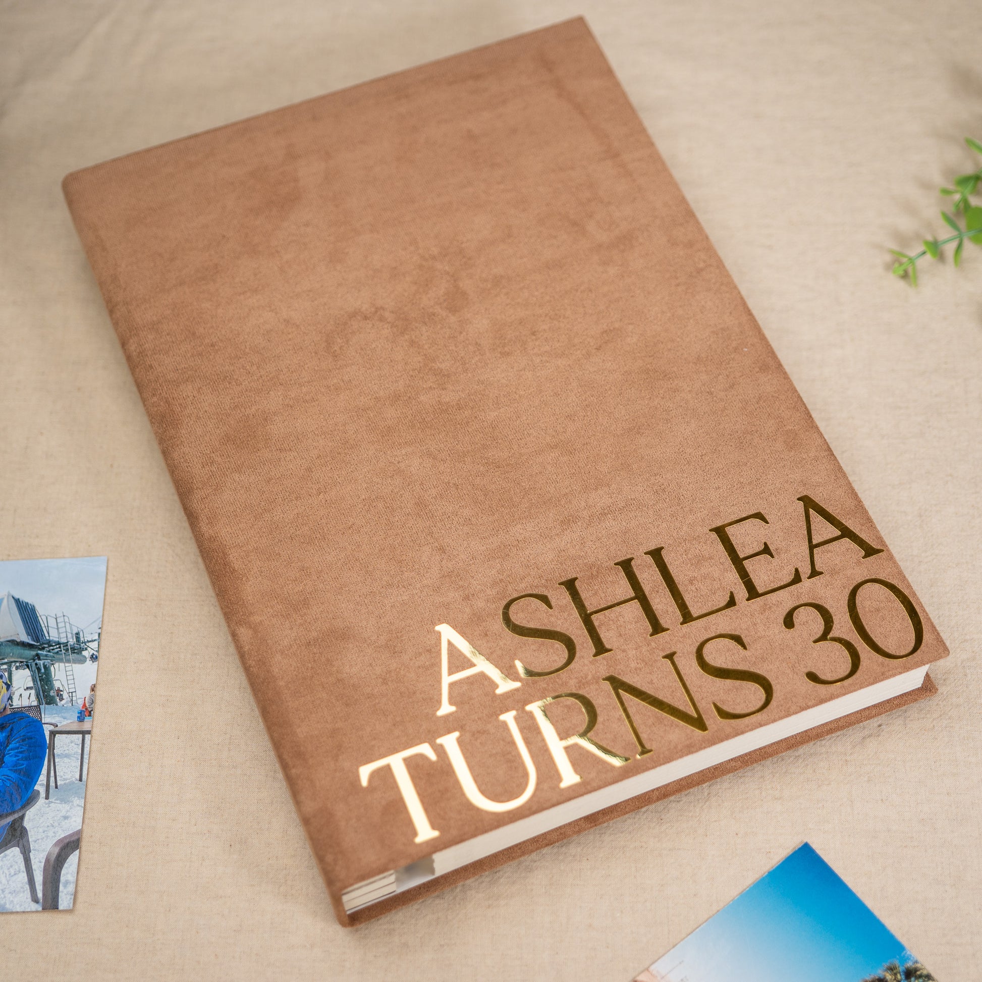 A personalized hardcover book or photo album with a textured tan cover and metallic gold foil text reading 'ASHLEA TURNS 30', photographed on a neutral fabric surface with small printed photos partially visible at the edges.