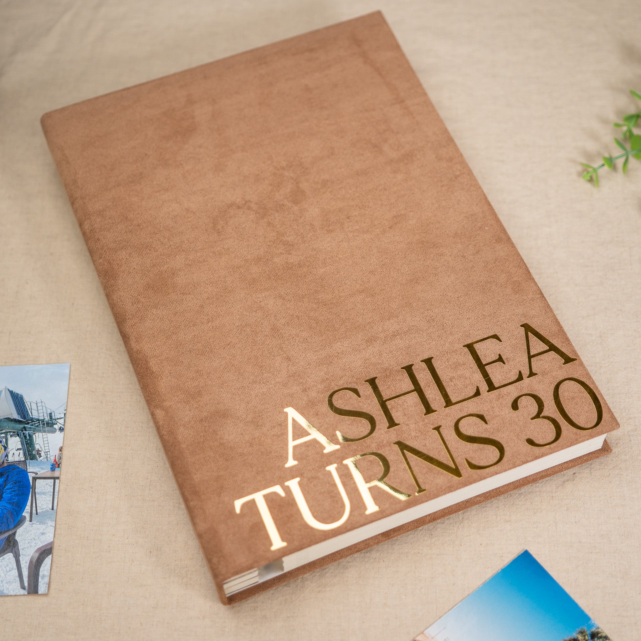A personalized hardcover book or photo album with a textured tan cover and metallic gold foil text reading 'ASHLEA TURNS 30', photographed on a neutral fabric surface with small printed photos partially visible at the edges.