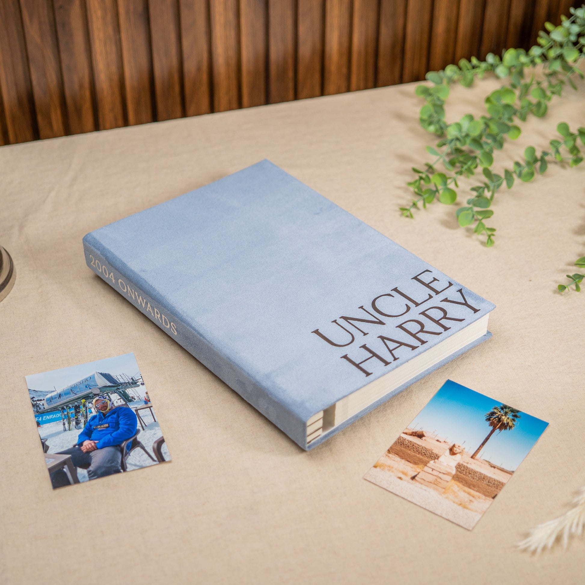 A hardcover book titled 'UNCLE HARRY'  lies on a beige tablecloth, styled with two small printed photos and green trailing leaves. The book cover is light blue with dark brown lettering, and the spine shows the author's name in matching dark lettering.