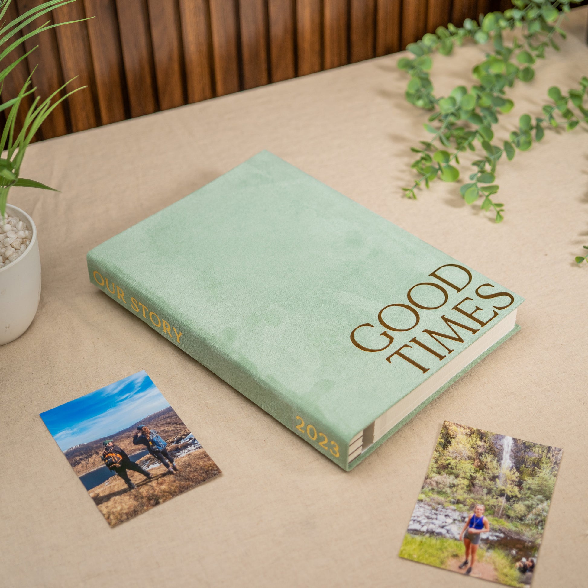 A mint green hardcover photo book titled 'OUR STORY' and 'GOOD TIMES 2023' lies on a beige tabletop, surrounded by small printed photos and green plants.